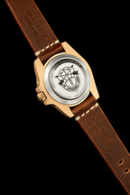 A close-up of a wristwatch with a gold-tone case and a brown leather strap, shown against a solid black background. The watch face is visible through the transparent back cover, featuring a black graphic design.