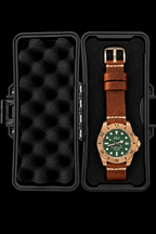 A wristwatch with a brown leather strap and green dial displayed inside an open black hard case with black foam padding on a black background.