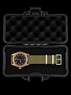 A wristwatch with a black dial, olive green NATO-style fabric strap, and rose gold case, displayed inside an open black hard case with black foam padding on a black background.